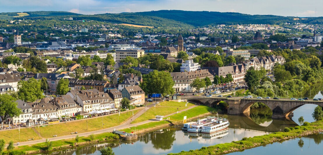 Blick auf Trier mit Mosel, historischen Gebäuden, Brücke und bewaldeten Hügeln im Hintergrund bei sonnigem Wetter.
