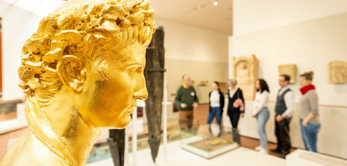 Goldene Büste mit lockigem Haar im Rheinischen Landesmuseum, im Hintergrund Besuchergruppe vor Ausstellungsstücken.