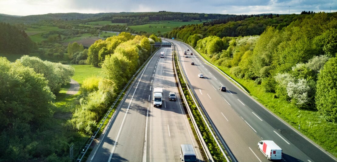 Autos auf einer mehrspurigen Autobahn in grüner Hügellandschaft bei Sonnenschein aus der Luftperspektive.