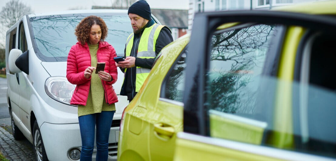 Eine Frau und ein Mann in gelber Warnweste im Austausch miteinander mit Smartphones in den Händen zwischen zwei geparkten Autos in einer Wohnsiedlung.