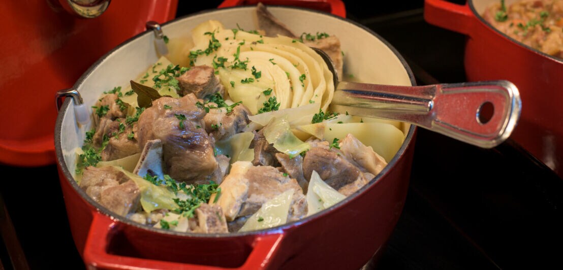 Roter Bräter mit Fårikål, bestehend aus Lammfleisch, Kohl und Kartoffeln, garniert mit Petersilie, daneben ein weiterer roter Topf mit Reisgericht.