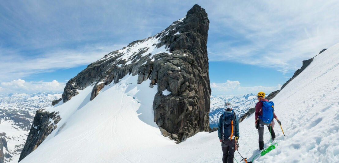 Zwei Bergsteiger mit Helmen und Rucksäcken stehen auf schneebedecktem Grat in Jotunheimen, Blick auf steilen Felsgipfel und umliegende Berglandschaft.