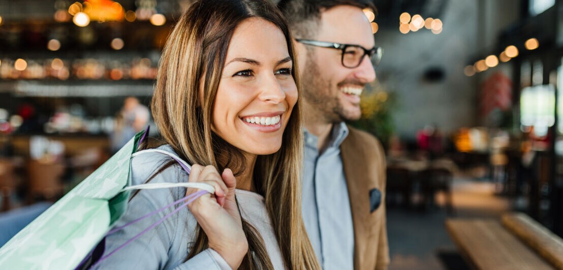 Eine Frau hält eine Tasche über der Schulter, neben ihr steht ein Mann, beide lächeln.