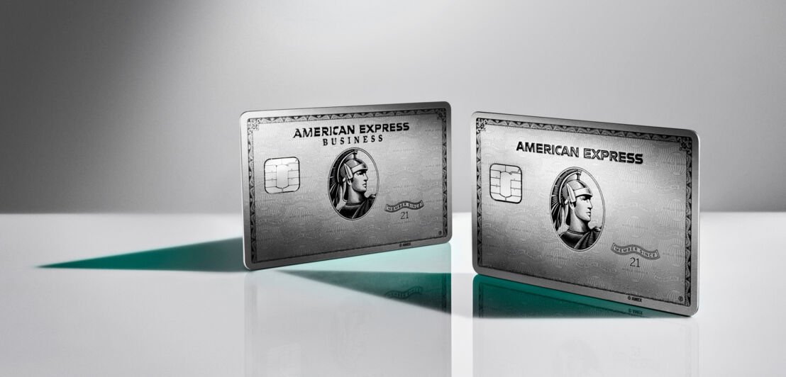 Die American Express Business Platinum Card neben der Platinum Plus Card, aufgestellt auf einer glänzenden Fläche vor grauem Hintergrund.