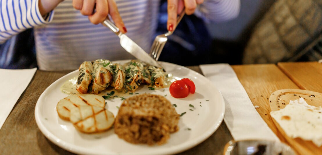 Hände einer Frau, die mit Messer und Gabel ein Gericht von einem weißen Teller isst.