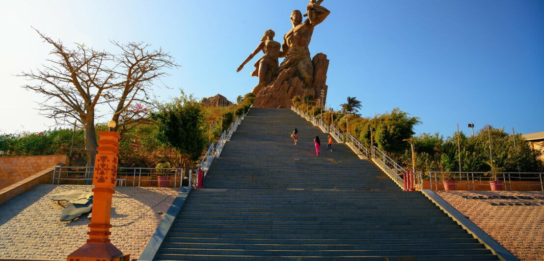 Personen auf einer pyramidenförmigen Treppe eines Denkmals mit großer Bronzestatue unter blauem Himmel.