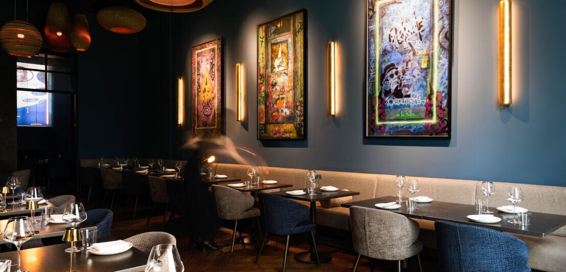 Modern eingerichteter Gastraum mit gedeckten Tischen, Stühlen in Grau und Blau, beleuchteten Wandbildern und warmem Licht an den Wänden.