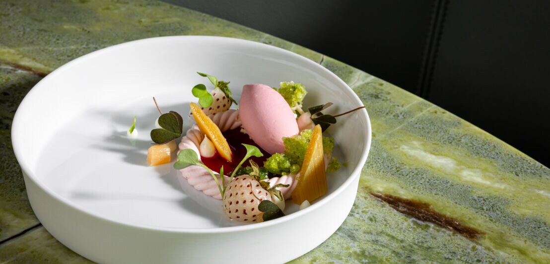 Weißer Teller mit rosa Sorbet, Babymais, weißen Erdbeeren und grünen Kräutern auf grün marmoriertem Tisch.