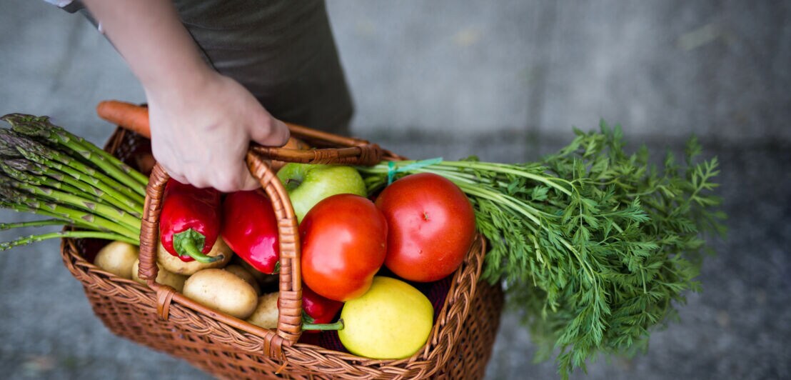 Hand hält geflochtenen Korb mit Biogemüse und Obst, darunter Tomaten, Karotten, Spargel, Paprika, Kartoffeln und ein Apfel.
