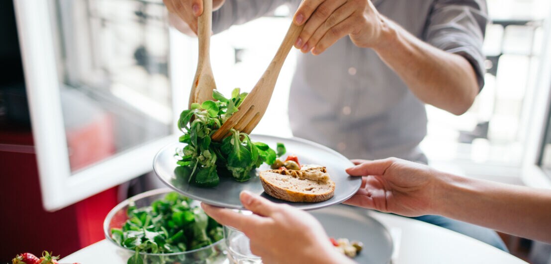Personen servieren frischen grünen Salat mit Holzbesteck auf Teller mit Brot und Gemüse, auf dem Tisch Schüssel mit Salat und Schale mit Erdbeeren.