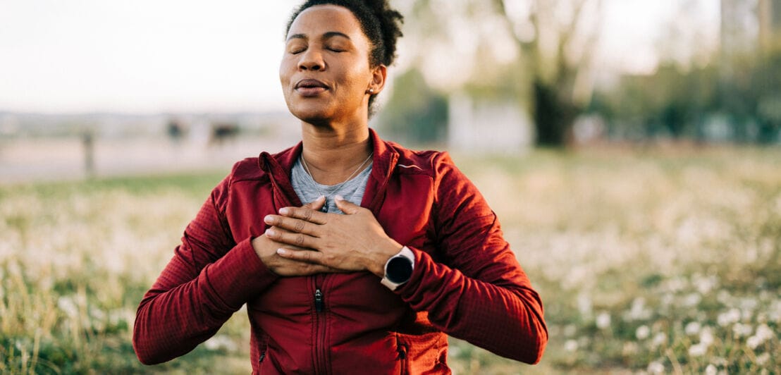Eine Person sitzt im Freien auf dem Gras, trägt einen roten Hoodie und hat die Hände auf der Brust. Im Hintergrund sind Bäume und ein verschwommener Park sichtbar.