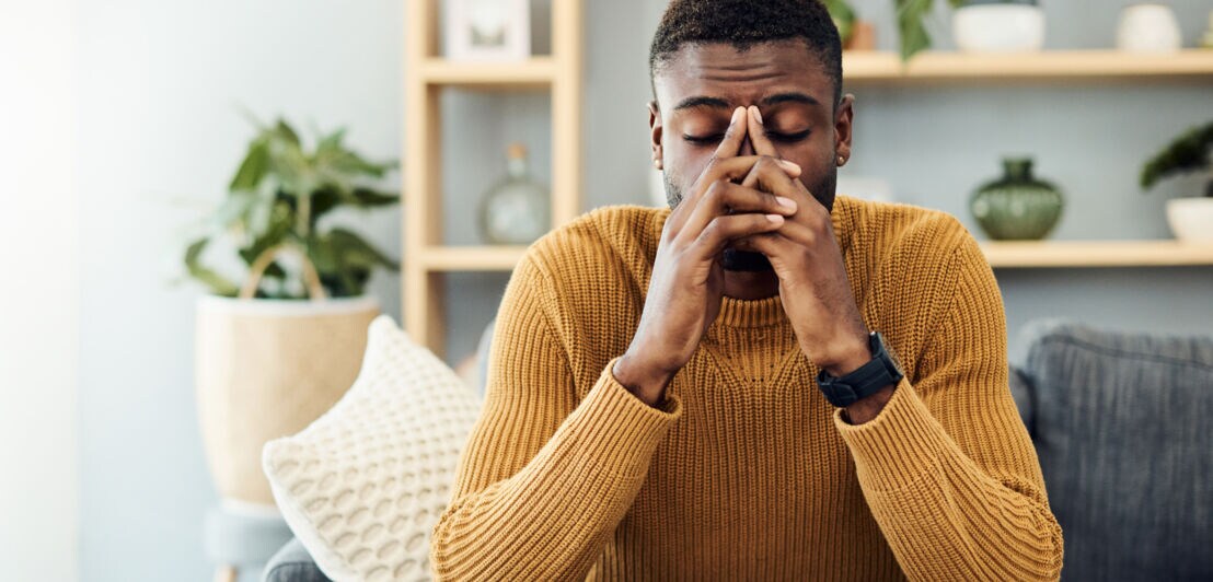 Ein Mann mit kurzem, dunklem Haar sitzt auf einem grauen Sofa, trägt einen orangefarbenen Pullover und schaut auf einen Laptop. Pflanzen und Regale im Hintergrund.
