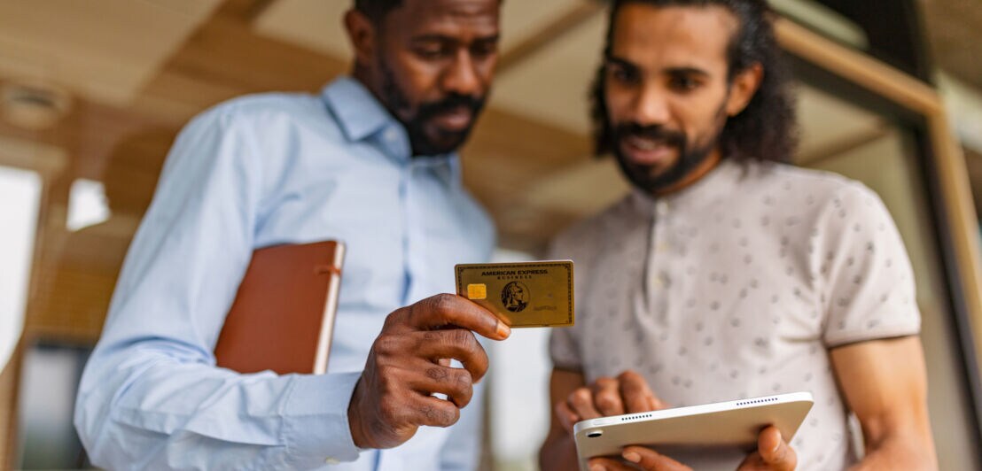 Zwei Männer schauen gemeinsam auf ein Tablet und eine goldene Kreditkarte von American Express in ihren Händen in einem Innenraum.