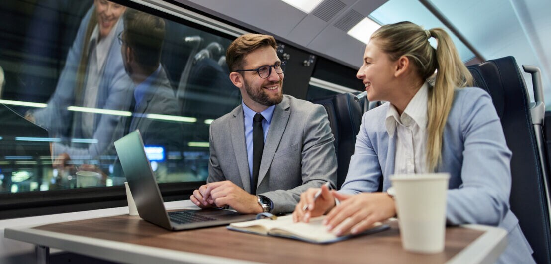 Ein Mann und eine Frau in Anzügen sitzen an einem Tisch in einem Zugabteil und unterhalten sich gut gelaunt vor einem Laptop.