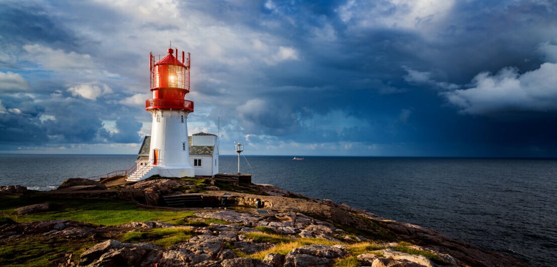 Roter und weißer Leuchtturm auf felsiger Küste unter dramatischem bewölktem Himmel am Meer in Norwegen.