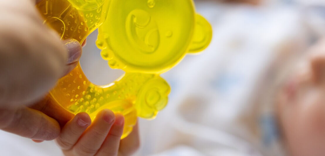 Hand hält gelbes, transparentes Baby-Beißspielzeug, das von einem Baby mit kleinen Fingern gehalten wird.