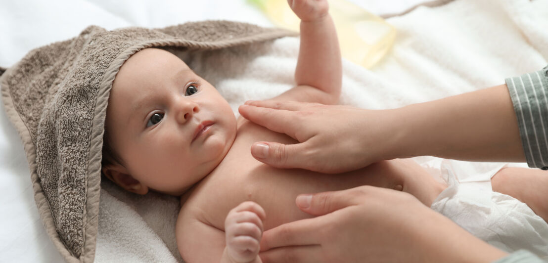 Baby liegt auf einer weißen Decke, teilweise mit braunem Handtuch bedeckt, während zwei Hände die Brust einreiben.