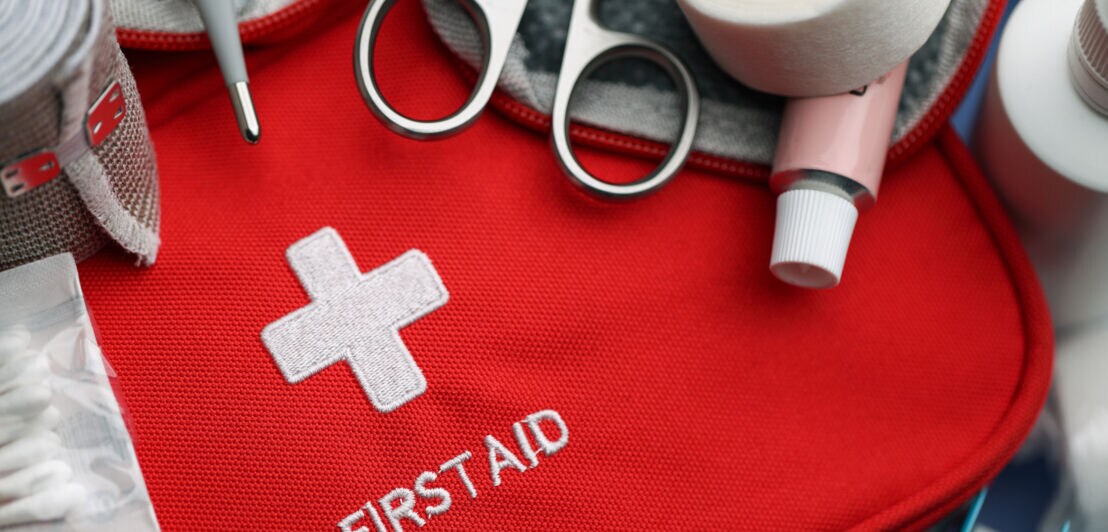 Roter Erste-Hilfe-Kasten mit einem weißen Kreuz und dem Text 'FIRST AID'. Umgeben von Scheren, Binden, Watte und verschiedenen medizinischen Utensilien.