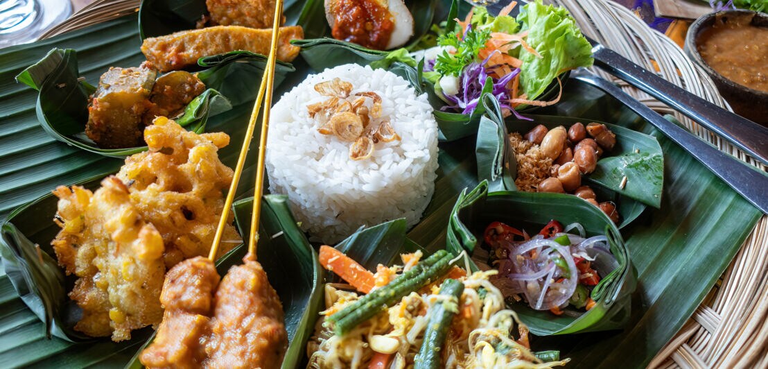 in runder Korb mit verschiedenen indonesischen Gerichten, darunter Reis, Gemüse, frittiertes Essen und Erdnüsse, serviert auf Bananenblättern.
