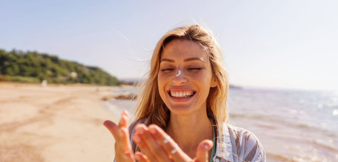 Eine lachende Frau mit Sonnencreme auf Händen und Gesicht an einem Strand.