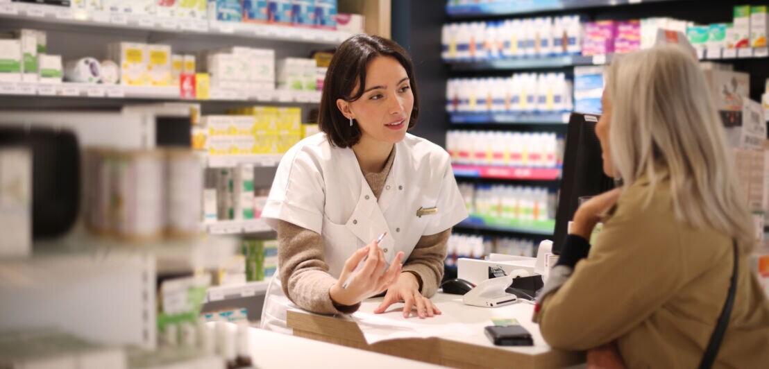 Eine Frau im weißen Kittel hinter einer Verkaufstheke spricht mit einer Kundin in einer Apotheke.