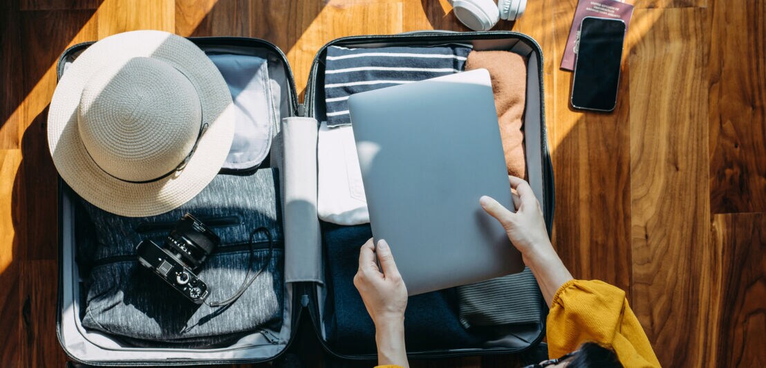 Offener Koffer mit Sonnenhut, Kamera, Kleidung, Laptop, weißen Kopfhörern und Smartphone auf Holzboden, Person legt Laptop in Koffer.