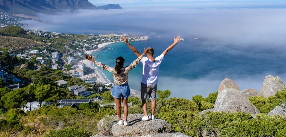 Zwei Personen stehen auf einem Felsen mit erhobenen Armen und blicken auf eine Küstenlandschaft mit Meer, Häusern und Bergen unter blauem Himmel.