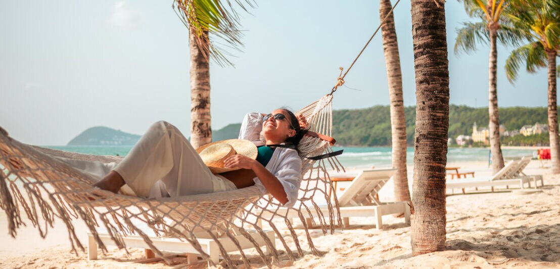 Eine Frau mit Sonnenbrille und Sonnenhut liegt entspannt in einer Hängematte an einem Strand in Vietnam.