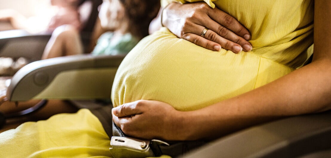 Nahaufnahme eines Schwangerschaftsbauch, umfasst von zwei Händen einer Frau im gelben Kleid auf einem Flugzeugsitz neben zwei Kindern.
