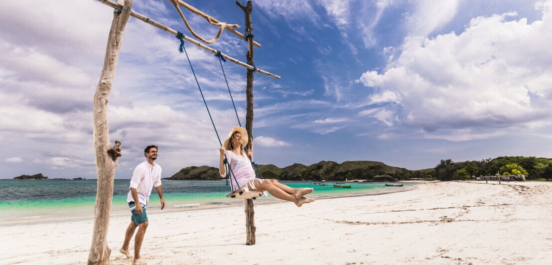 Ein Mann steht hinter einer schaukelnden schwangeren Frau auf einer Holzschaukel an einem weißen Sandstrand am türkisblauen Meer.