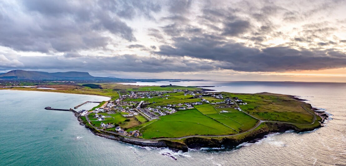 Luftaufnahme von einer grünen Halb-Insel mit kleinen Häuser, die von einem Meer umgeben ist.