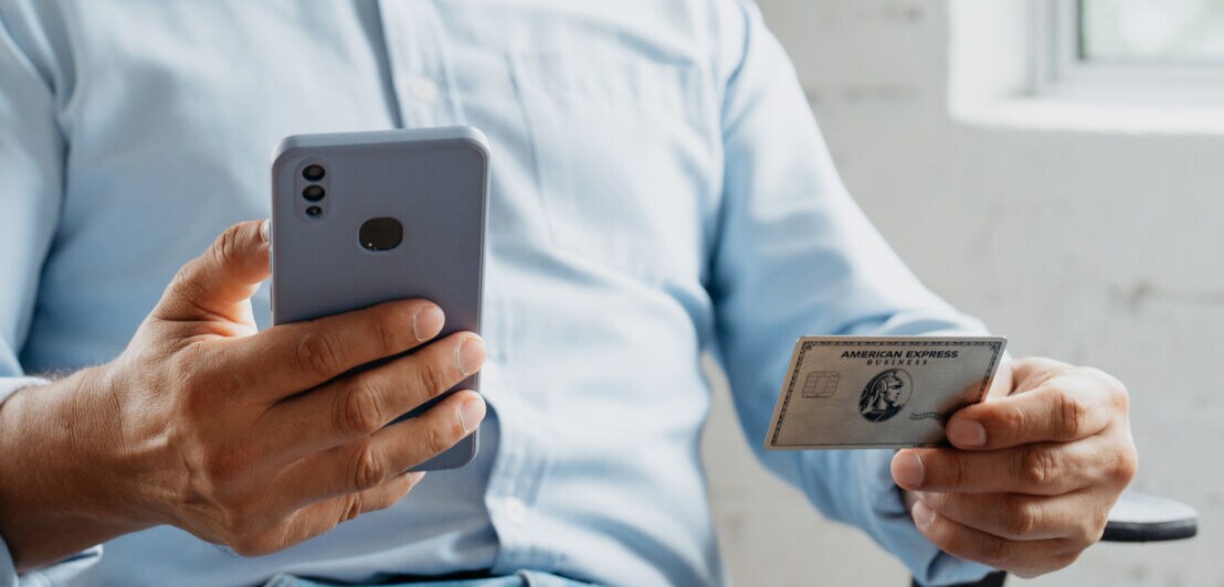 Ein Mann in hellblauem Hemd hält ein Smartphone und eine Business Platinum Card von American Express in einer, ein Smartphone in der anderen Hand.