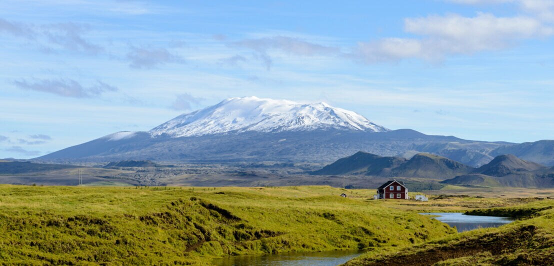 Einsames Haus in weitläufiger grüner Landschaft, mit dem schneebedeckten Vulkan Hekla im Hintergrund.