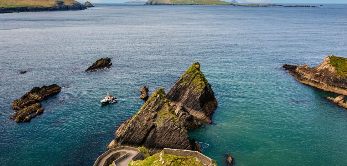 Blick von oben auf eine Küstenlandschaft mit einem geschwungenen Steinweg, Felsen im Wasser und Inseln im Hintergrund.