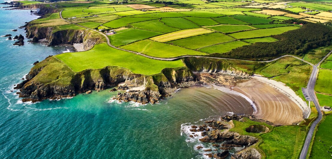 Luftaufnahme der grünen Felder und Küstenklippen an der Copper Coast in County Waterford, Irland, mit Straßen und Bergen im Hintergrund.