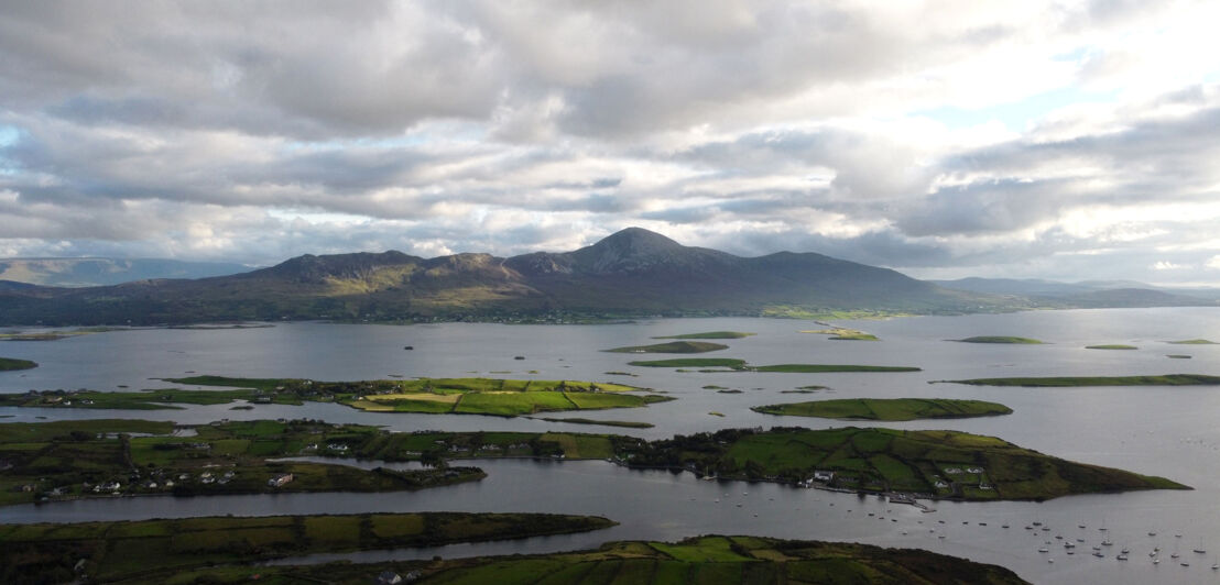 Vor einer Küste liegen etliche kleine, grüne Inseln im Wasser, im Hintergrund ragt ein großer Berg, umgeben von Wolken, empor.