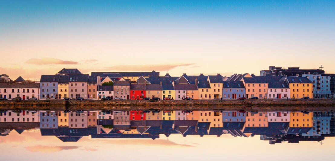 Etliche bunte, kleine Häuser stehen an einem Wasser, die Häuser spiegeln sich in der Wasseroberfläche.