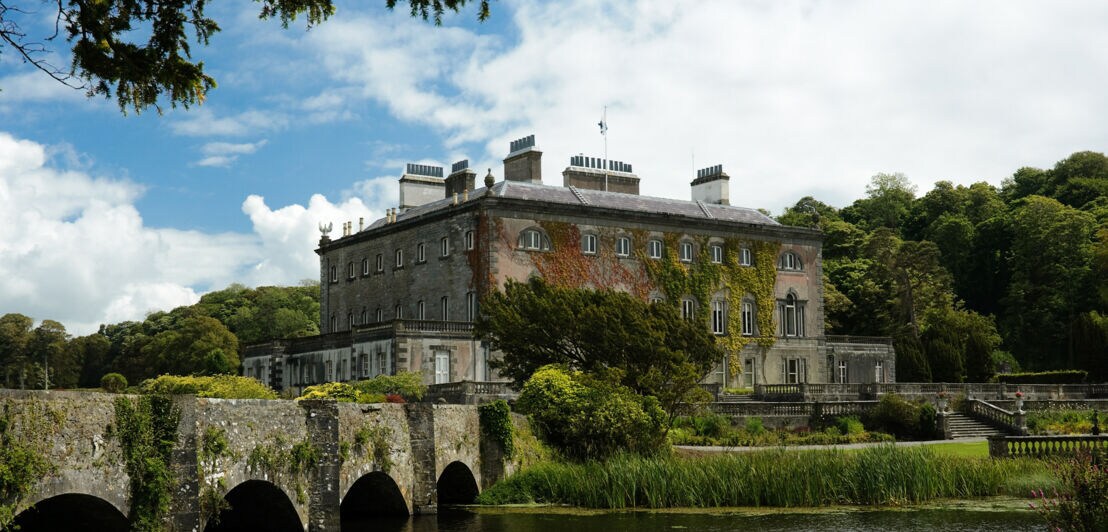 Vor einem großen, rechteckigen Haus mit vielen kleinen Fenstern liegt ein See mit einer stabilen Steinbrücke.