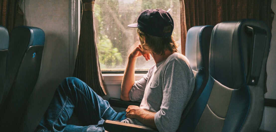 Ein Mann mit einer Baseballmütze sitzt in einem Zug und schaut nachdenklich aus dem Fenster. Er trägt ein graues Oberteil und Jeans.