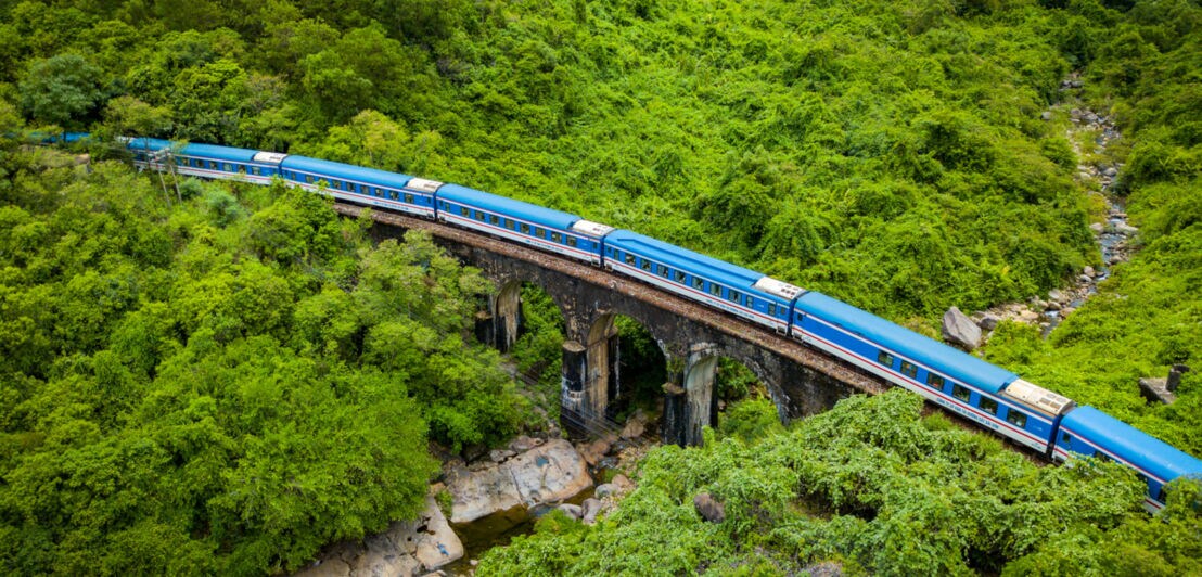 Ein blauer Zug fährt über eine Brücke aus Stein, umgeben von dichtem, grünem Wald und Felsen. Im Vordergrund ist ein Flussbett sichtbar.