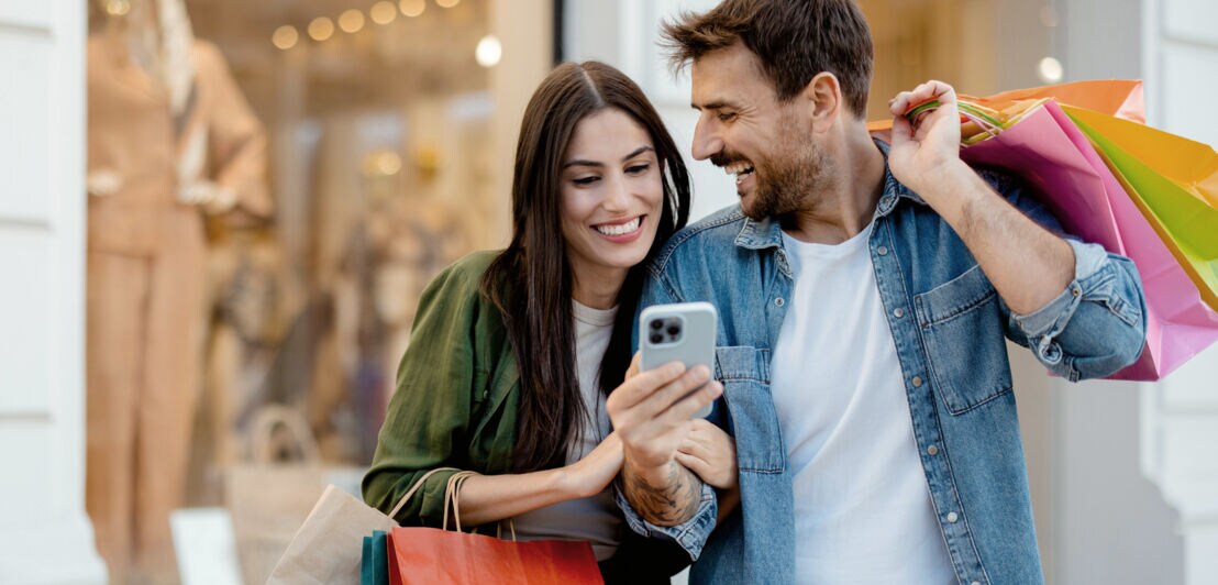Eine Frau und ein Mann stehen eingehakt und gut gelaunt vor einem Modegeschäft mit bunten Einkaufstüten in den Händen, vor ihnen ein hochgehaltenes Smartphone in seiner Hand.