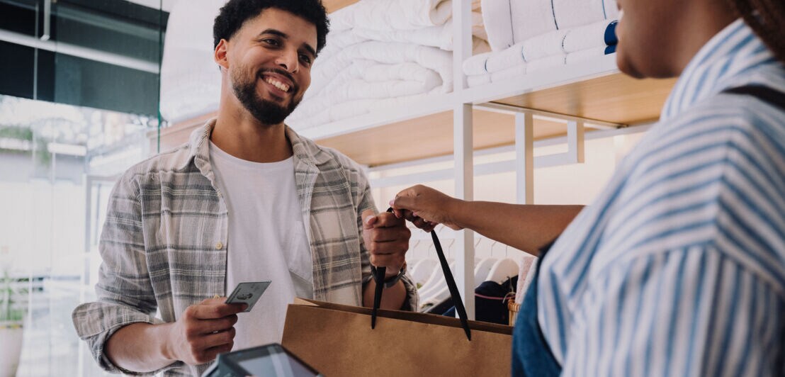 Ein Mann nimmt lächelnd eine Einkaufstüte von einer Verkäuferin in einem Geschäft entgegen, dabei hält er eine silberne Kreditkarte von American Express in der Hand.