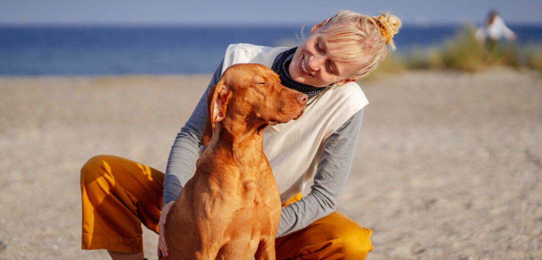 Eine Frau kniet hinter einem großen Hund mit goldfarbenem Fell und schaut ihn lächelnd an auf einem breiten Sandstrand bei Sonnenschein.