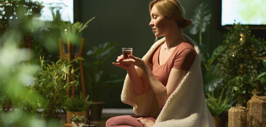 Frau sitzt im Schneidersitz auf dem Boden, hält eine kleine Tasse, trägt rosa Sportkleidung und eine weiße Decke über den Schultern, umgeben von Pflanzen.