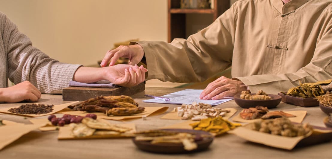 Zwei Personen sitzen an einem Tisch mit verschiedenen getrockneten Kräutern und Wurzeln, eine Person hält das Handgelenk der anderen zur Pulsmessung.