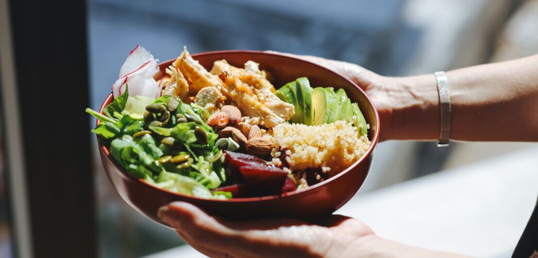 Schale mit gemischtem Salat, Avocadoscheiben, Quinoa, Nüssen und gebratenem Hähnchen, gehalten von zwei Händen.