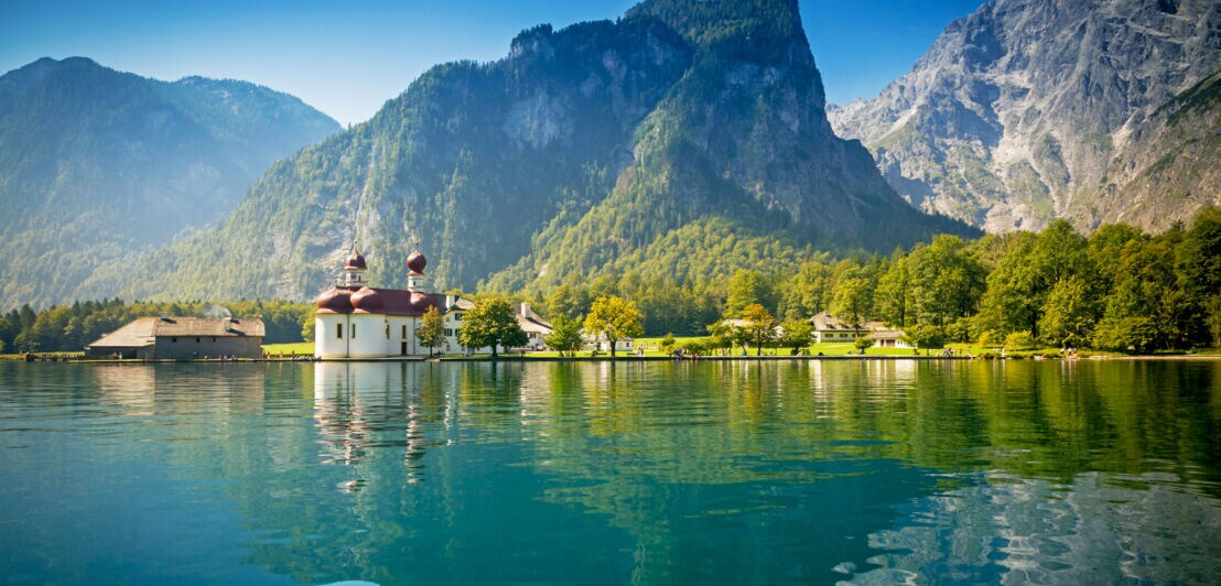 Kirche mit Zwiebeltürmen am Ufer eines klaren Sees, umgeben von bewaldeten Bergen unter blauem Himmel.