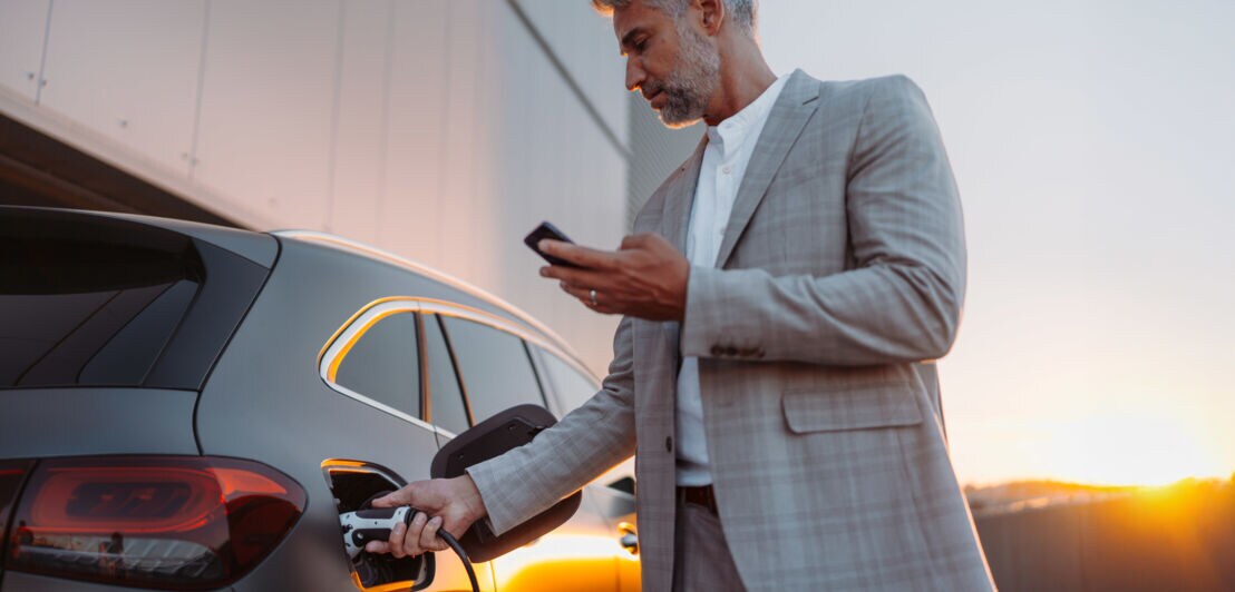 Ein Mann im grauen Anzug und mit einem Smartphone in der Hand steckt den Stecker einer E-Ladesäule in ein Auto.