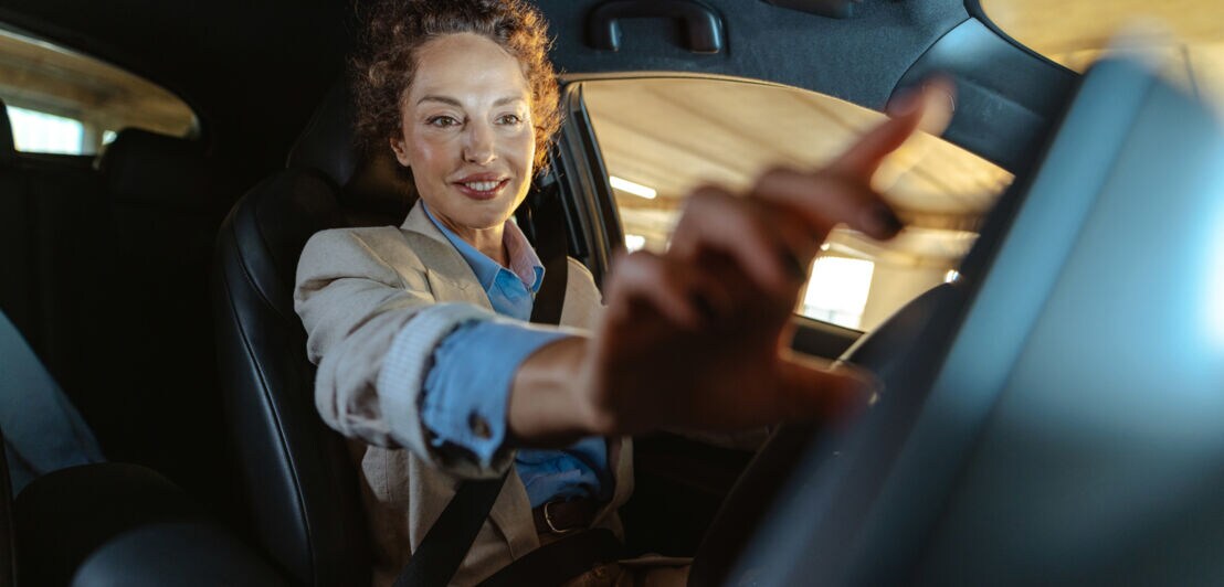 Eine lächelnde Frau sitzt am Steuer eines Autos und streckt ihre Hand nach dem Armaturenbrett aus.
