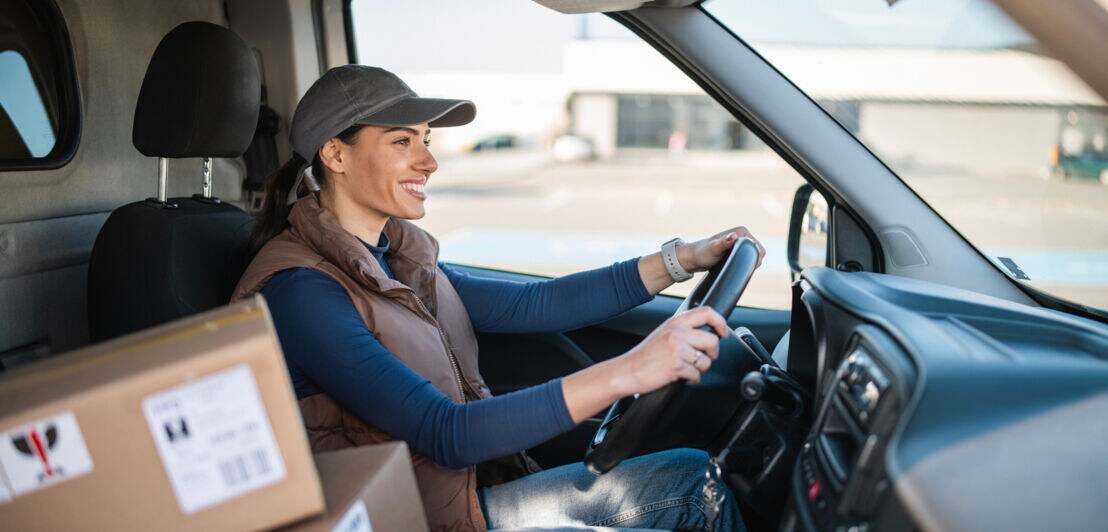 Eine lächelnde Frau mit Basecap sitzt am Steuer eines Transporters. Auf dem Beifahrersitz liegen gestapelte Kartons.